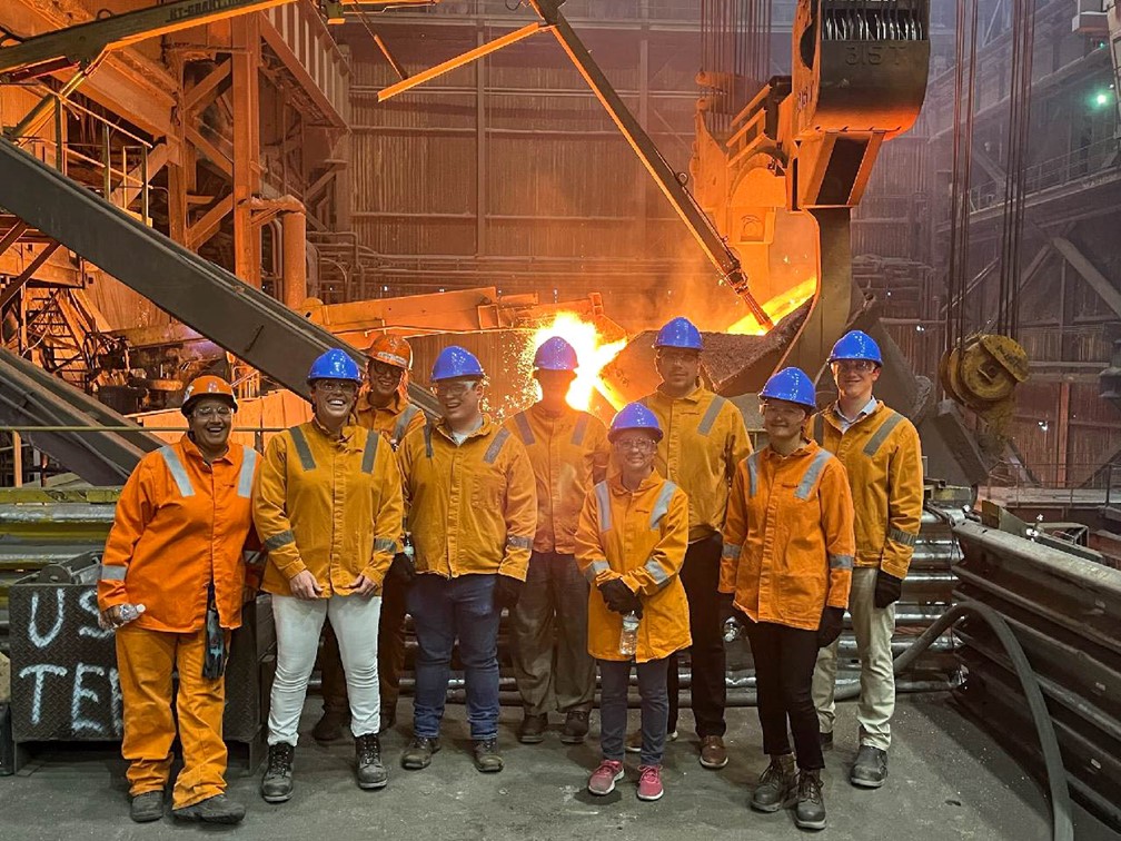 The USTR team stands alongside steel workers from Edgar Thompson steel plant as molten steel is poured behind them.